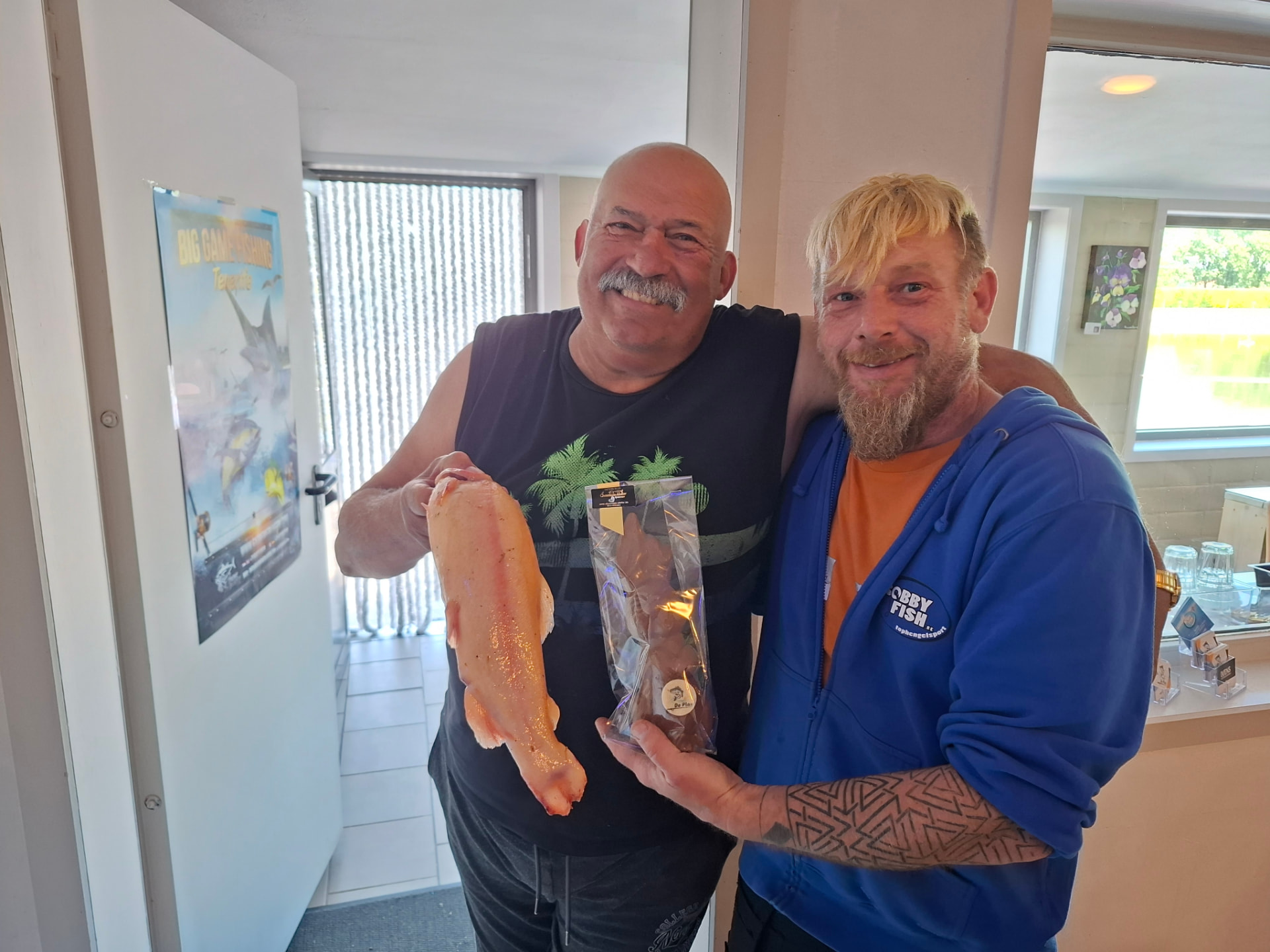 Twee mannen lachen naast elkaar, één houdt een forel vast